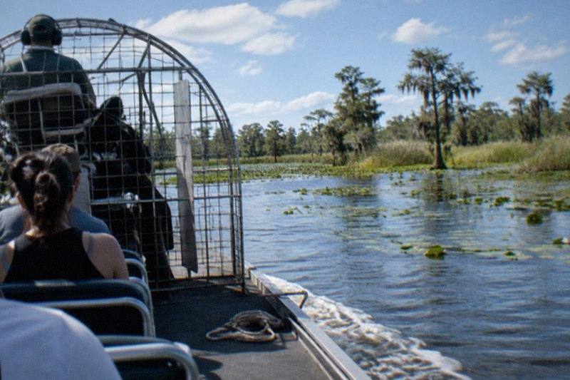 An airboat crossing bright open water at midday, cypress trees and sawgrass along the far edge of the marsh, no people, no logos, no readable text.