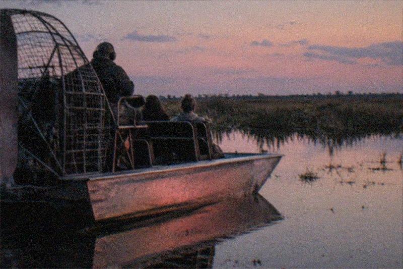 An airboat drifting across still water as the sun sets, warm light washing the surface and marsh grass dark against the shore, no people, no logos, no readable text.