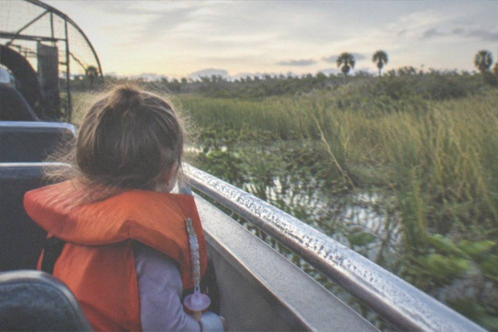 A child in a life jacket watching tall marsh grass pass from an airboat bench seat