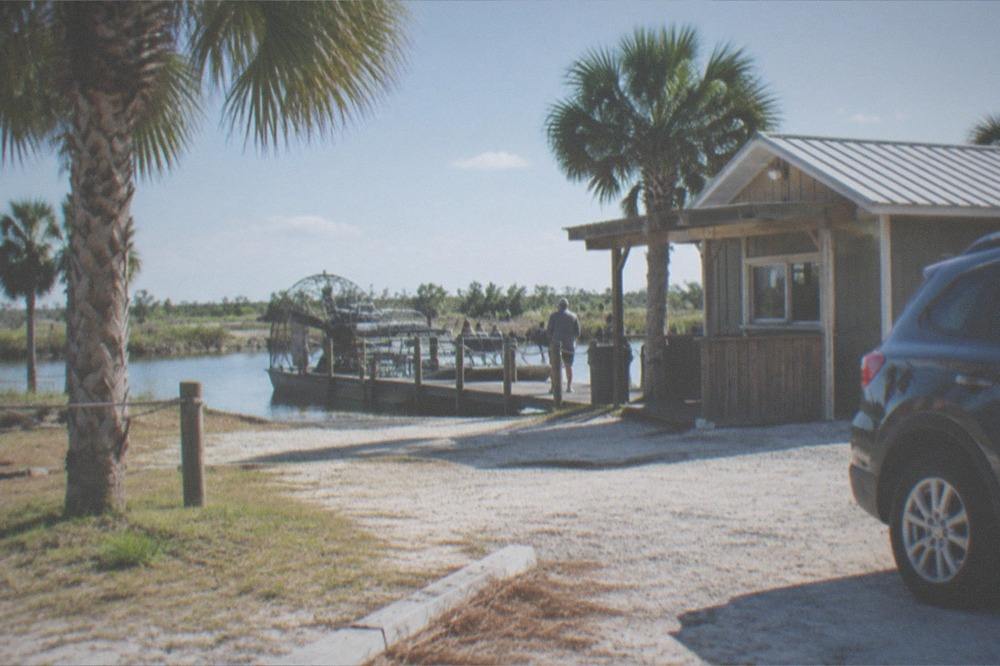 A parked car beside a dockside ticket booth at a Kissimmee airboat launch on a bright morning
