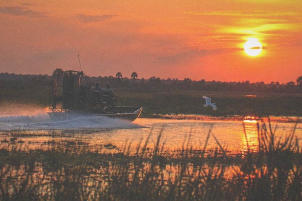 An airboat cruising through open marsh water with the sky glowing orange at sunset