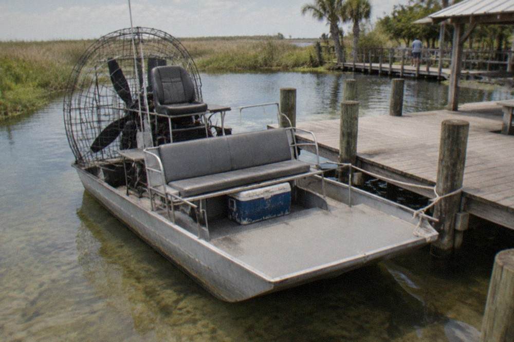 A wide flat-bottomed airboat parked at a wooden dock on a shallow Florida lake, tall marsh grass behind it