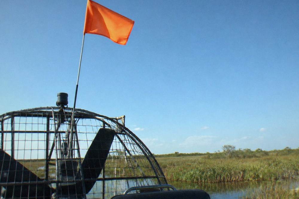 A tall international-orange safety flag mounted on a mast above an airboat, seen against a bright sky over a marsh