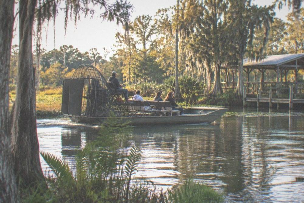 A small airboat carrying a few passengers across a quiet marsh channel
