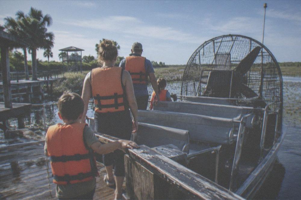 A family in life vests boarding an airboat at a wooden dock in morning light