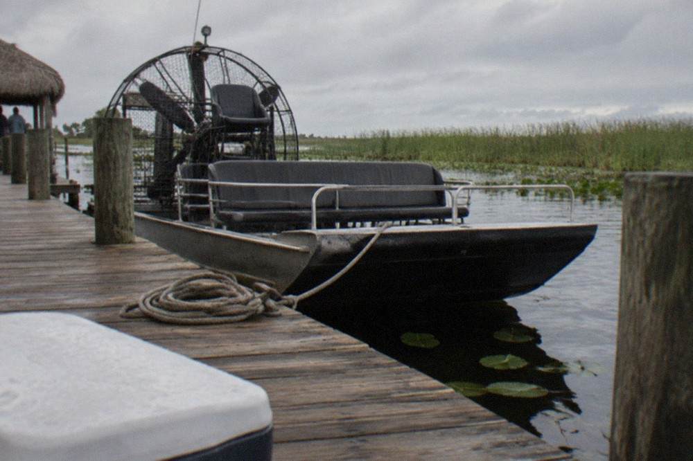 A small airboat parked at a wooden dock beside a marsh, reeds and open water in the background