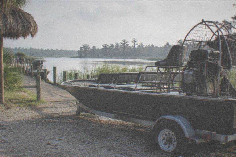 A parked airboat on a gravel launch ramp at a Central Florida lake shore, reeds and distant treeline visible