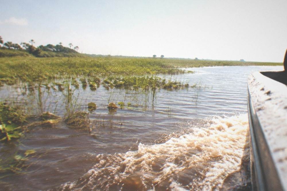 A low airboat cutting a wake across shallow brown marsh water, grass flats stretching to the horizon