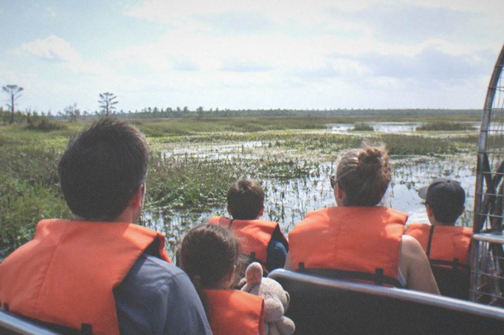 A family in life vests seated together on a small airboat moving through open marsh
