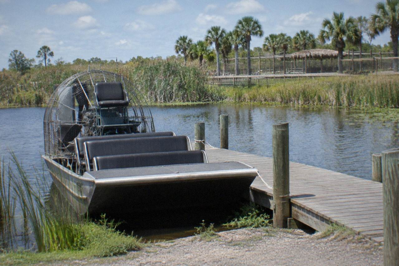 Combo-tour image. Airboat tied to a rustic dock on a Central Florida lake at midday, cypress and reed beds behind it, a wildlife park entrance in the distance near Orlando, Florida, United States, no people, no logos, no readable text.