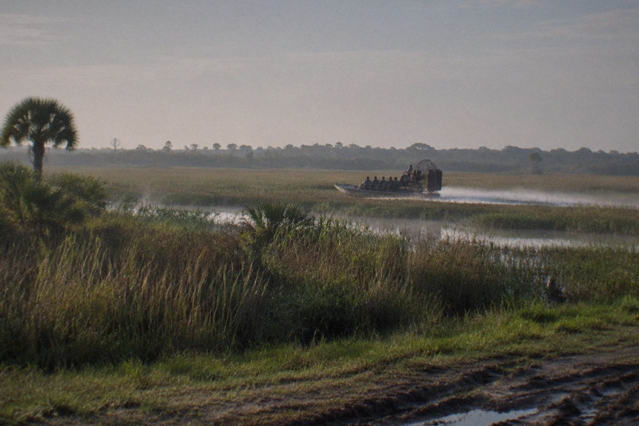 A wide view of shallow Florida wetland with an airboat cutting a wake through reeds, flat grassland running to the distant treeline, empty of people and signage.