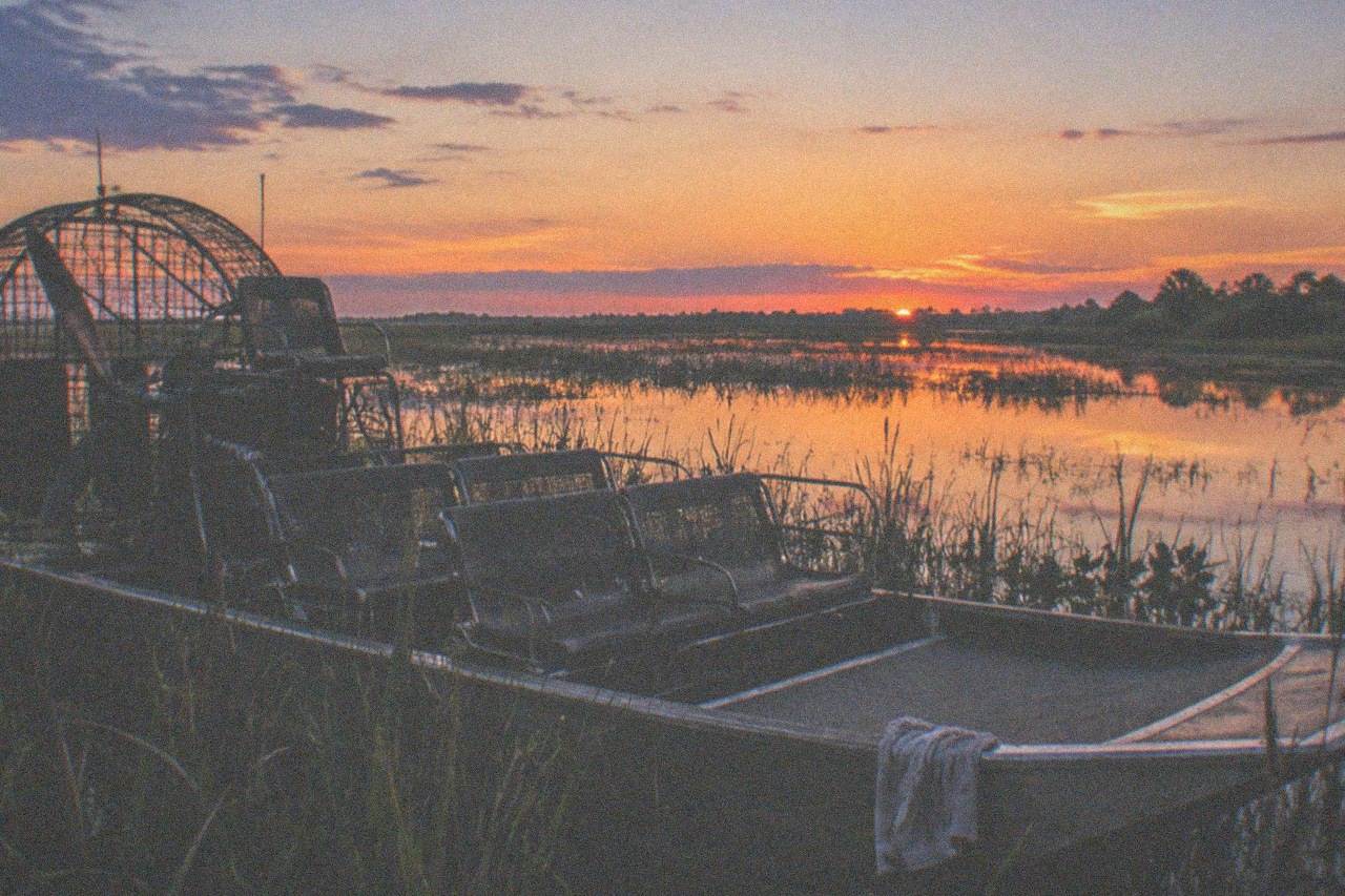 An airboat idling among cattails while dusk settles over a Central Florida lake, the sky shifting from amber to violet and the calm water mirroring the last light, empty seats, no people, no logos, no readable text.